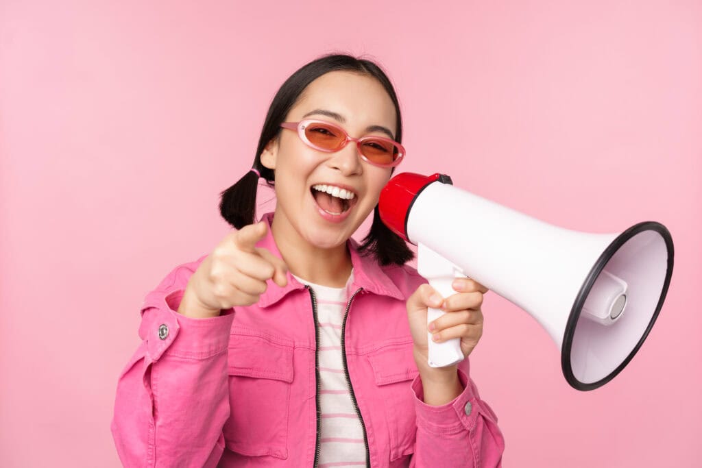 Smiling woman in pink jacket and sunglasses holds a megaphone and points forward against a pink background. | Tsunami Reach