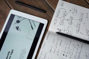 Tablet displaying a presentation beside a notebook with handwritten notes and a black pen on a wooden table. | Tsunami Reach