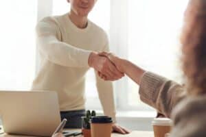 Two people shaking hands across a desk with coffee cups and a laptop in a bright office setting. | Tsunami Reach