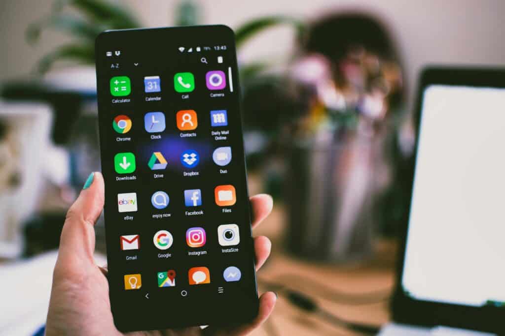 A hand holds a smartphone displaying various app icons, with a blurred desk and laptop in the background. | Tsunami Reach