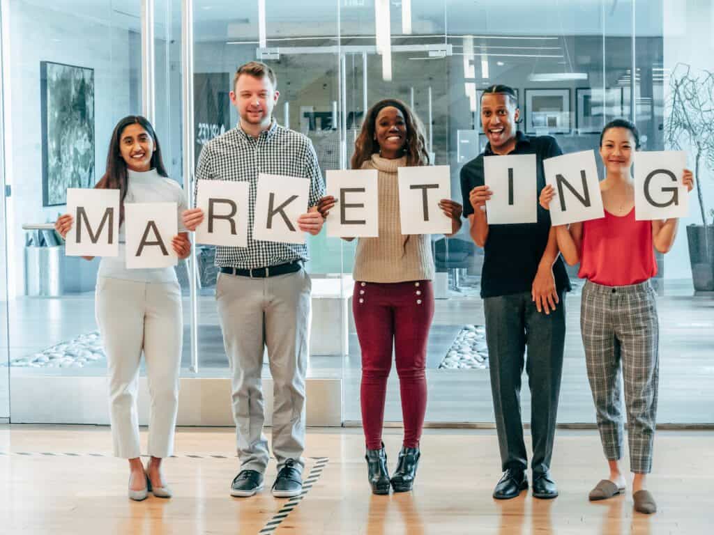 Five people standing in a row, each holding a letter to spell "MARKETING" inside an office space. | Tsunami Reach