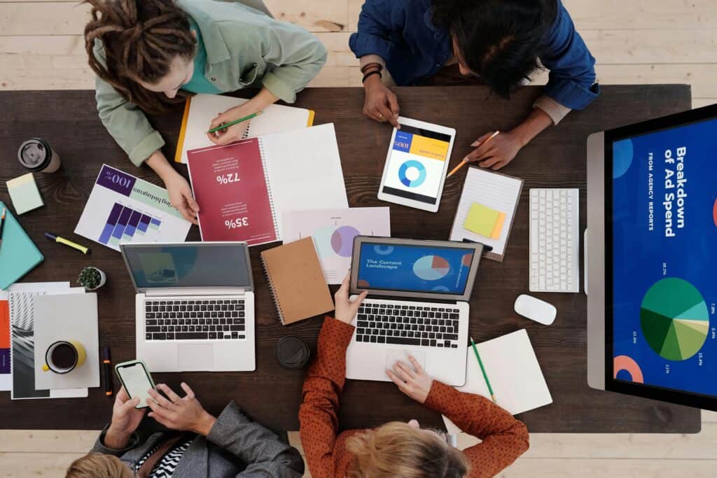 Four people work at a table with laptops, tablets, notebooks, and charts displaying graphs and data analysis. | Tsunami Reach