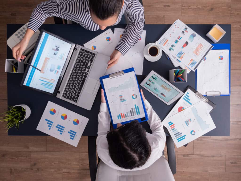 Two people at a desk analyzing charts and graphs on paper, a laptop, and a tablet, with coffee and office supplies nearby. | Tsunami Reach
