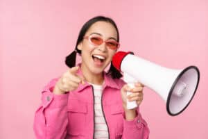 Smiling woman in pink jacket and sunglasses holding a megaphone and pointing at the camera, on a pink background. | Tsunami Reach