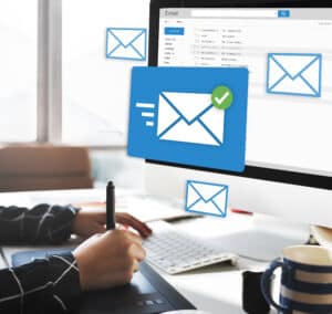 Person using tablet at desk with email icons and a computer screen showing an email inbox, signifying online communication. | Tsunami Reach