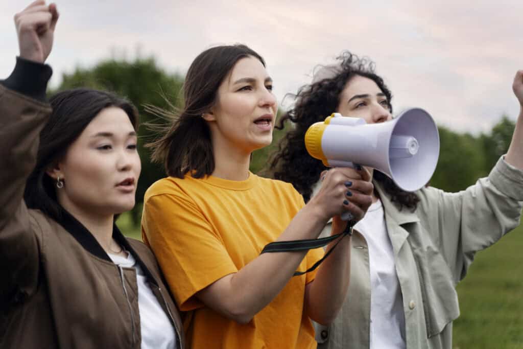 Three women standing outdoors, one holding a megaphone, all appearing to protest or chant with raised fists. | Tsunami Reach