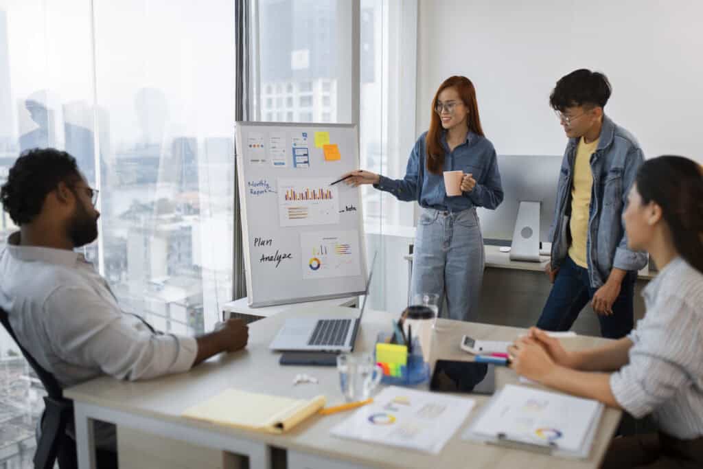 Four people in a modern office discuss charts on a whiteboard during a business meeting. | Tsunami Reach