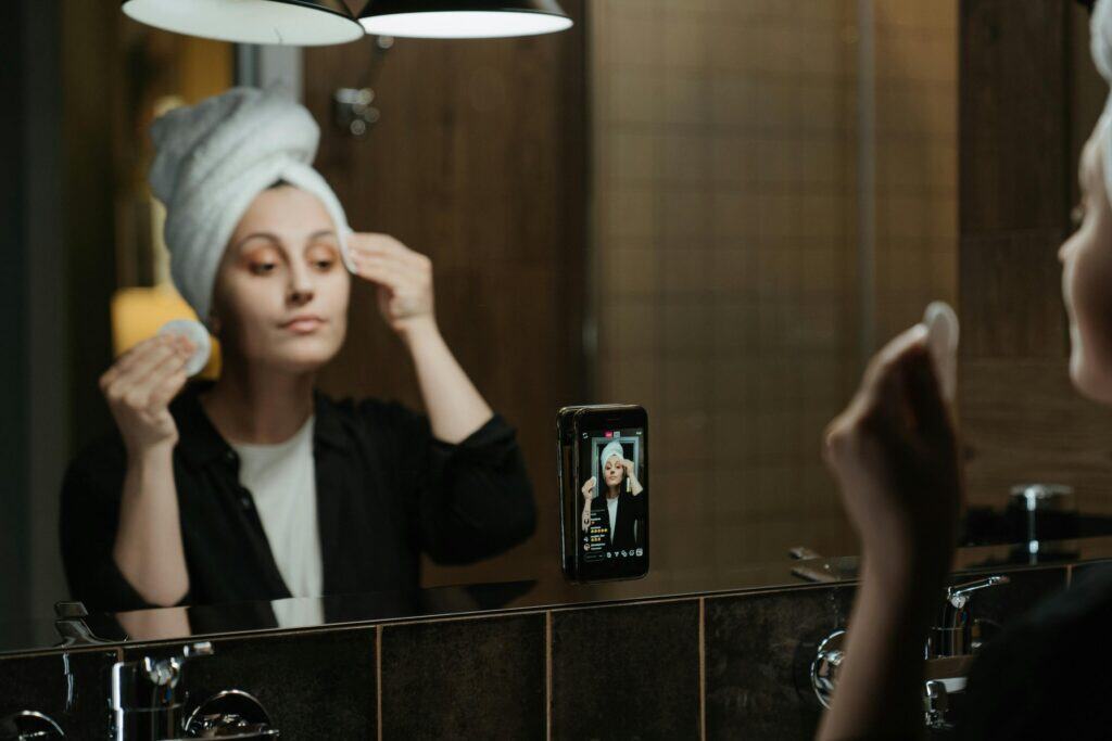 Woman with a towel on her head applies skincare in front of a mirror while recording herself on a smartphone. | Tsunami Reach