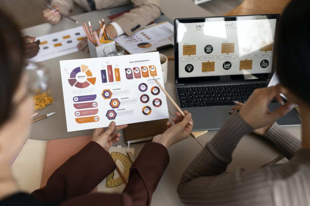 Two people review printed charts and infographics at a desk with a laptop displaying a presentation. | Tsunami Reach