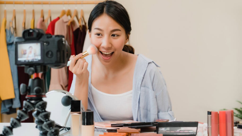 Woman applies makeup with a brush while filming herself, surrounded by cosmetics and clothes in the background. | Tsunami Reach