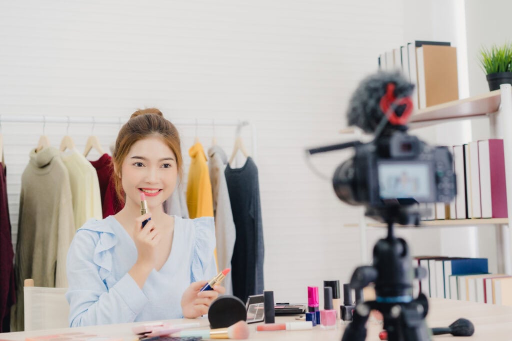 Woman recording a makeup tutorial, holding a lipstick in front of a camera, with cosmetics on the table. | Tsunami Reach