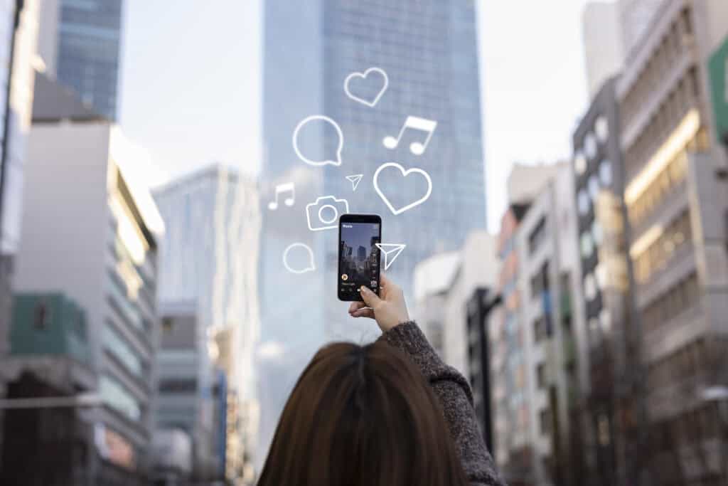 Person holding up a smartphone in a city, with social media icons like hearts and music notes drawn above the phone. | Tsunami Reach