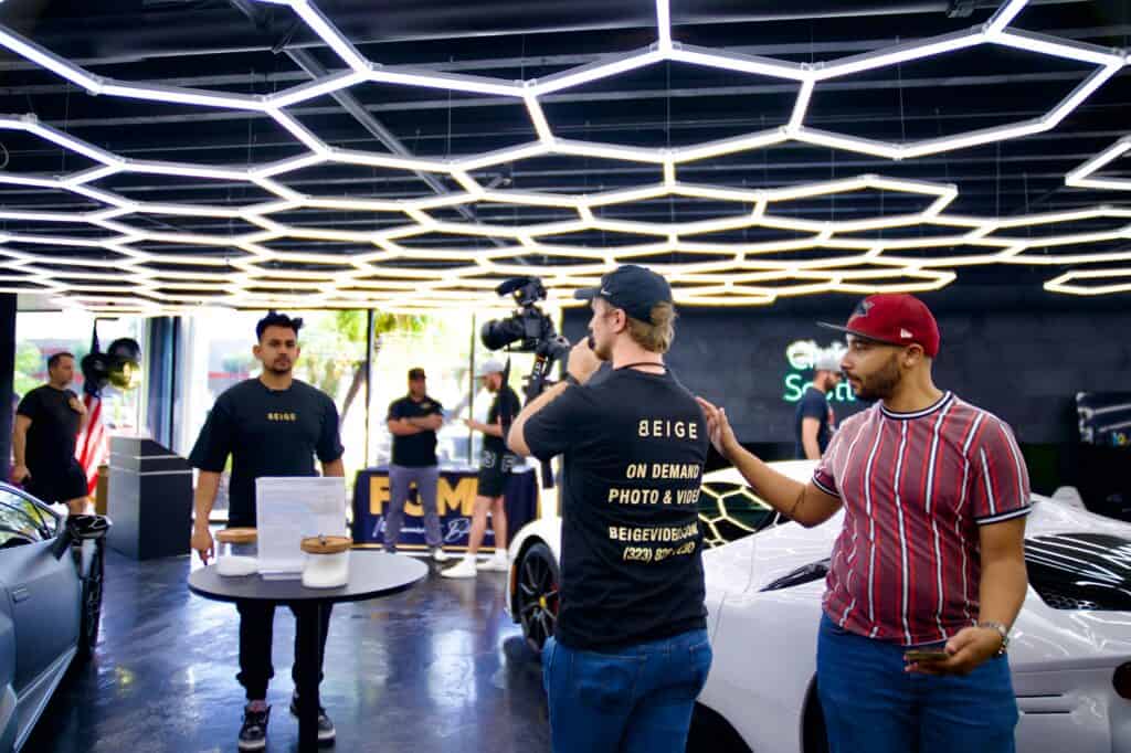 A cameraman films a man in black while another man in a red cap gestures in a modern showroom with luxury cars. | Tsunami Reach