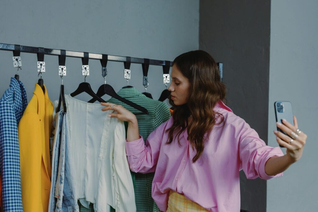 Woman in pink shirt takes a selfie while looking at clothes on a rack in a room. | Tsunami Reach