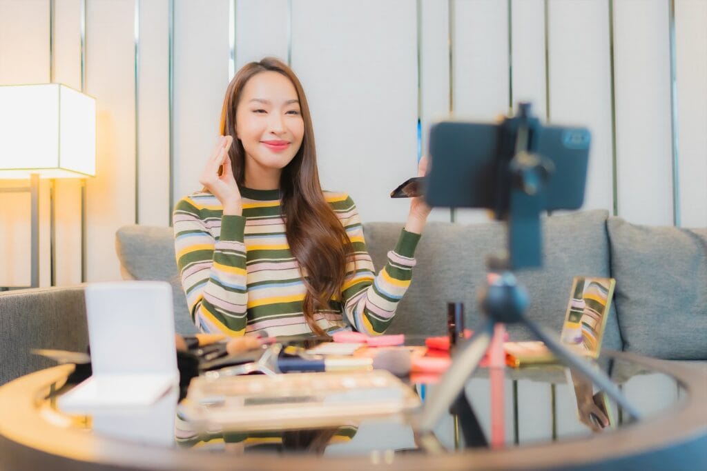 Woman recording a makeup tutorial at home, applying makeup with products on the table and a phone on a tripod. | Tsunami Reach