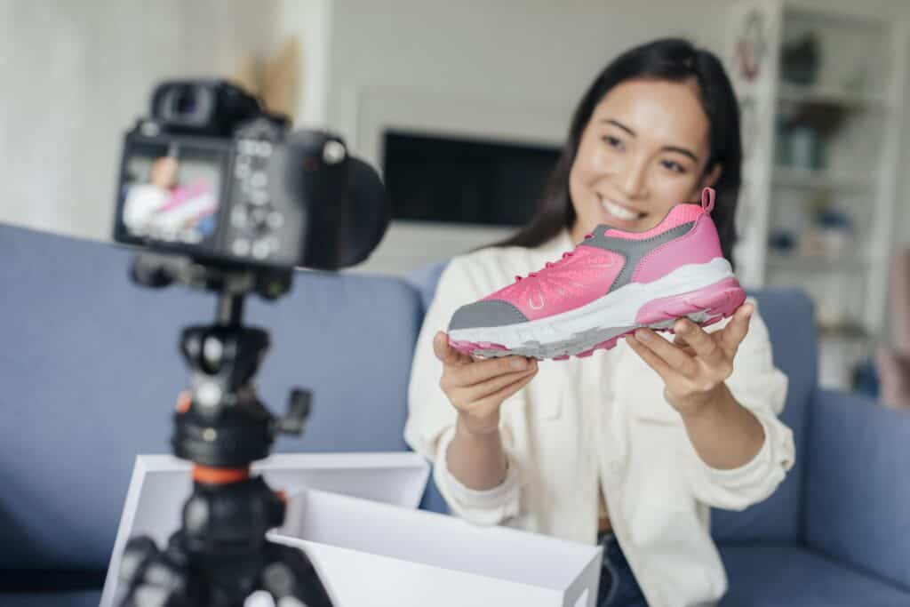 Smiling woman holds a pink sneaker and records a video with a camera on a tripod in her living room. | Tsunami Reach