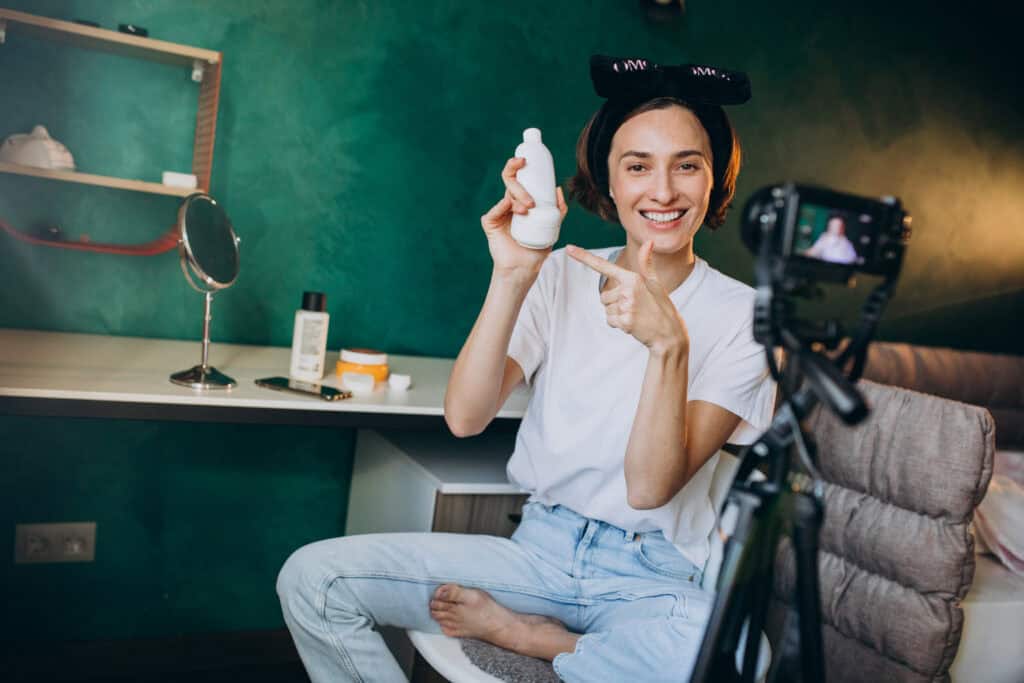 Smiling woman with hair rollers holds and points to a bottle while filming herself in a bedroom setup. | Tsunami Reach