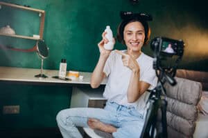 Smiling woman with hair rollers holds and points to a bottle while filming herself in a bedroom setup. | Tsunami Reach