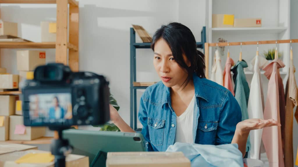 Woman filming herself while talking in a room with clothes and boxes, possibly creating content or doing a livestream. | Tsunami Reach
