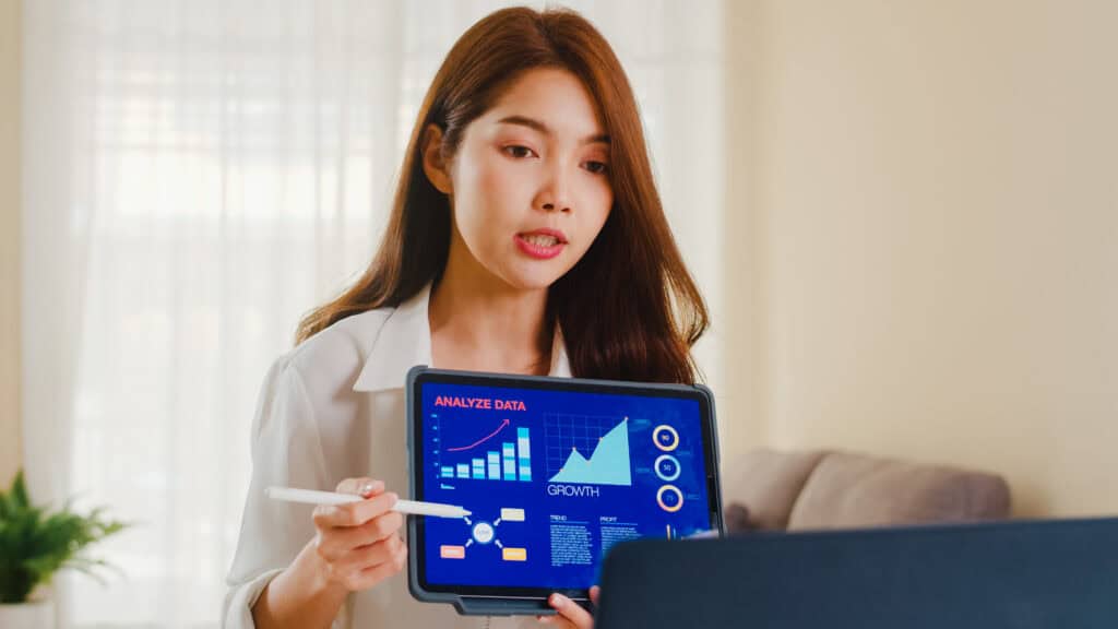 Woman holding a tablet showing data graphs during a video call, explaining analysis with a stylus in hand. | Tsunami Reach