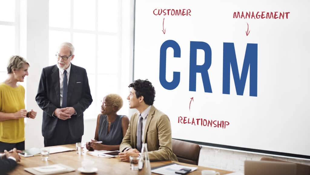 Four professionals discuss in a meeting room; a big screen shows "CRM" with arrows to "Customer," "Relationship," and "Management. | Tsunami Reach
