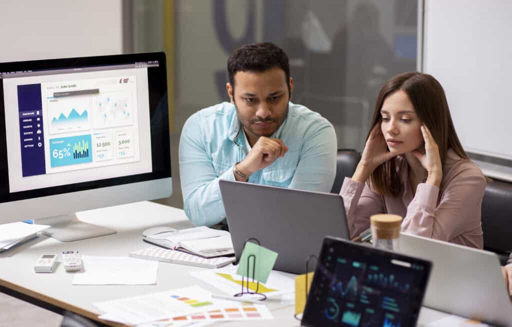 Two coworkers look stressed while working at a desk with laptops, charts, and graphs displayed on screens. | Tsunami Reach