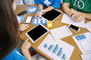 People working at a table with tablets, smartphones, charts, and social media icons, collaborating on a project. | Tsunami Reach