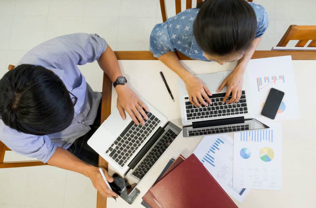 Two people work on laptops at a table with charts, papers, a phone, and folders spread out around them. | Tsunami Reach