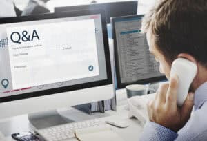 Man on phone at desk with a computer displaying a Q&A form and another screen showing a spreadsheet. | Tsunami Reach