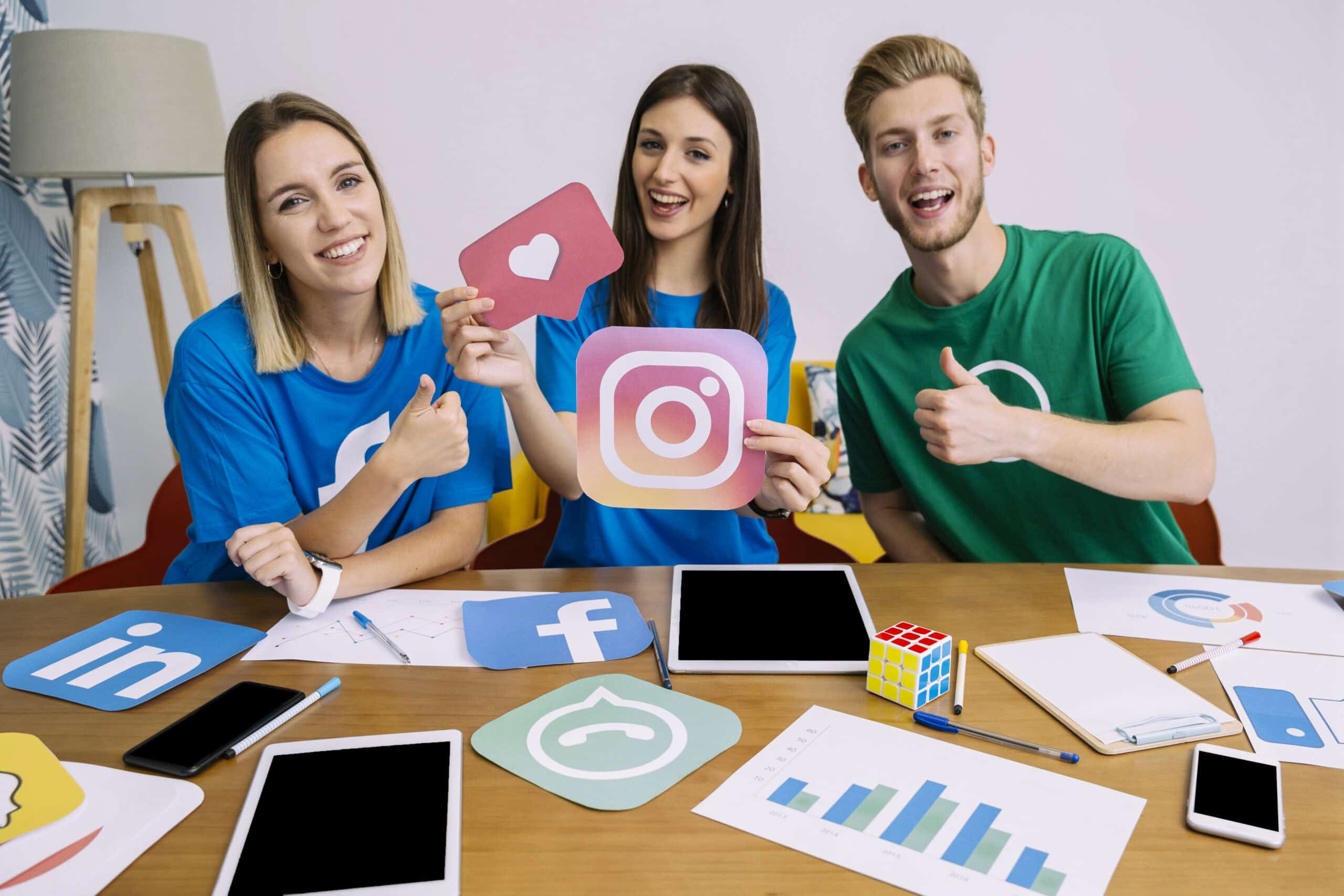 Three smiling people at a desk with social media icons, planning a marketing strategy with tablets, charts, and a Rubik’s cube. | Tsunami Reach
