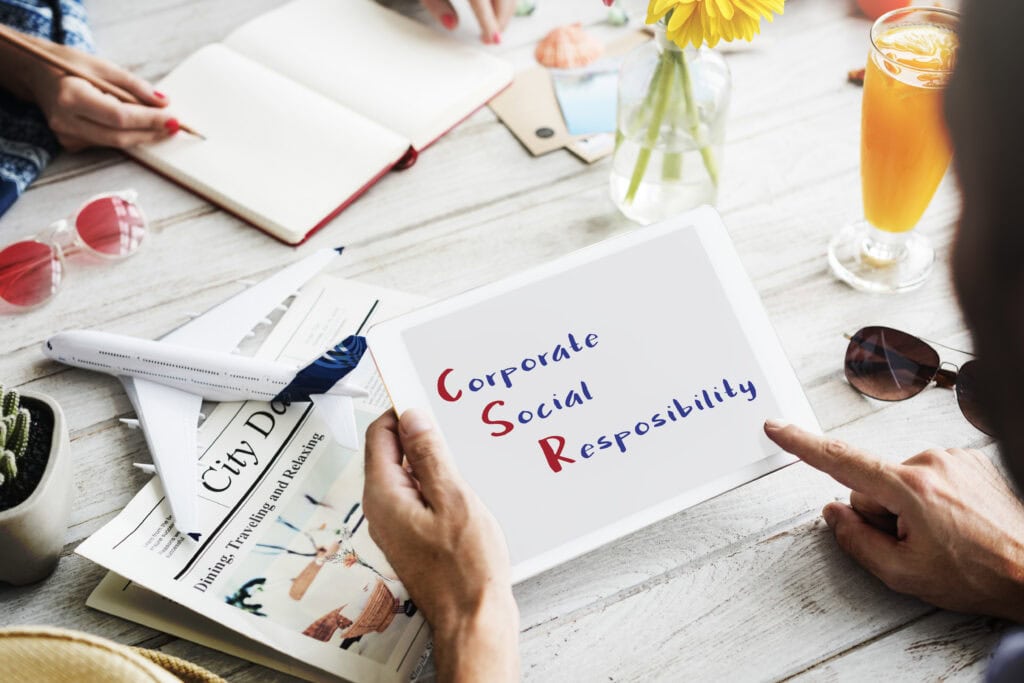 Person holding tablet with “Corporate Social Responsibility” text at a table with drinks, papers, and stationery. | Tsunami Reach