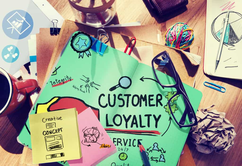 Notebook with “Customer Loyalty” written on it, surrounded by glasses, papers, sticky notes, and office supplies. | Tsunami Reach