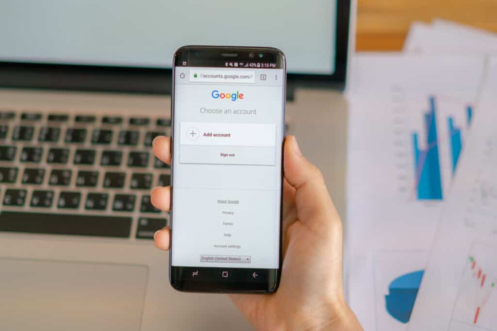 A hand holds a smartphone with a Google account login screen displayed, in front of a laptop and documents. | Tsunami Reach