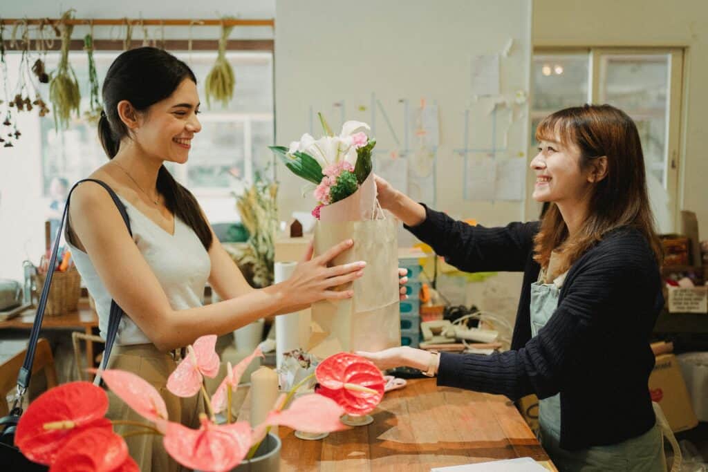 Two women smiling as one hands a bouquet of flowers in a paper bag to the other inside a flower shop. | Tsunami Reach