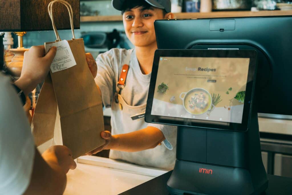 Smiling cashier hands a paper bag to a customer at a counter with a touchscreen register displaying soup recipes. | Tsunami Reach