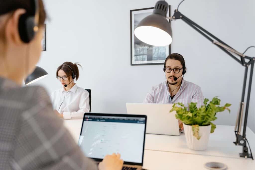 Three people with headsets work at laptops in a modern office with white walls and a desk lamp overhead. | Tsunami Reach