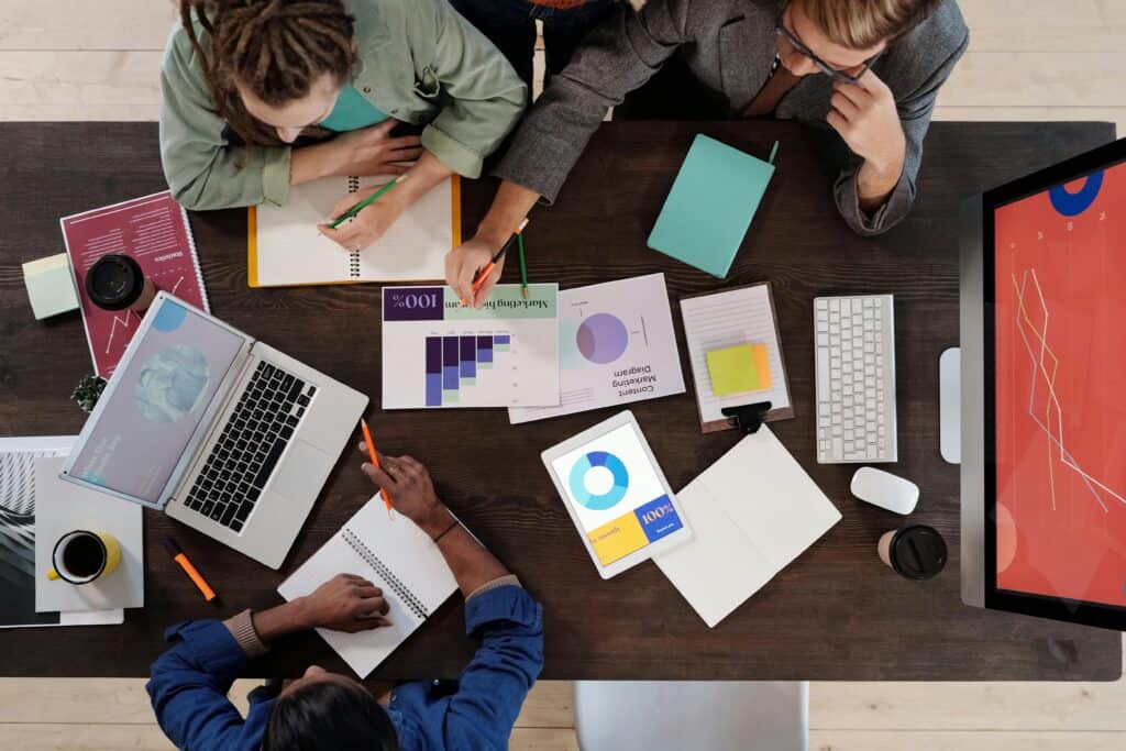 Three people collaborate at a desk with laptops, charts, notebooks, and graphs displayed on paper and a tablet. | Tsunami Reach
