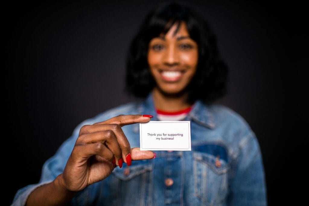 A smiling woman in a denim jacket holds a card that says, “Thank you for supporting my business!”. | Tsunami Reach
