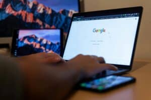 Person using a laptop with Google on screen, with a phone and two monitors in the background displaying mountains. | Tsunami Reach