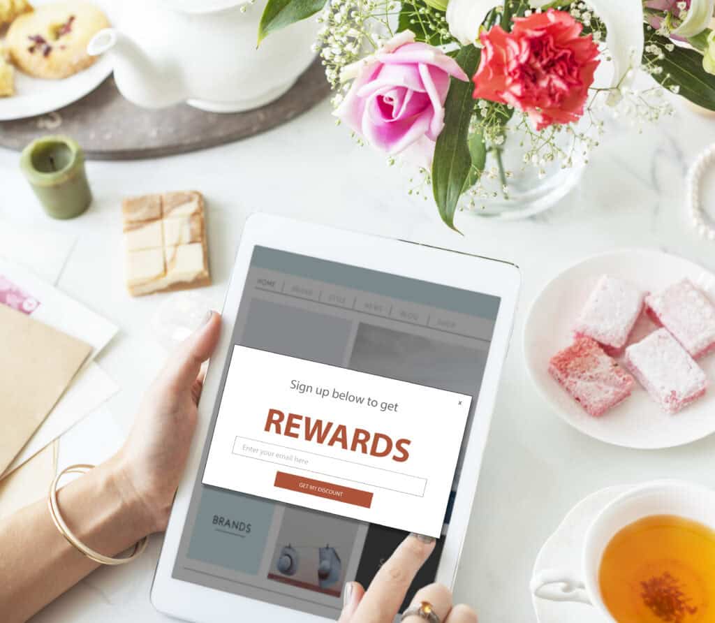 Person using a tablet with a rewards sign-up pop-up, surrounded by flowers, tea, and pastries on a table. | Tsunami Reach