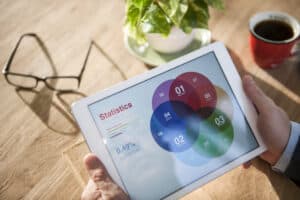 Person holding a tablet displaying colorful overlapping circle charts and statistics, with glasses, a plant, and coffee nearby. | Tsunami Reach