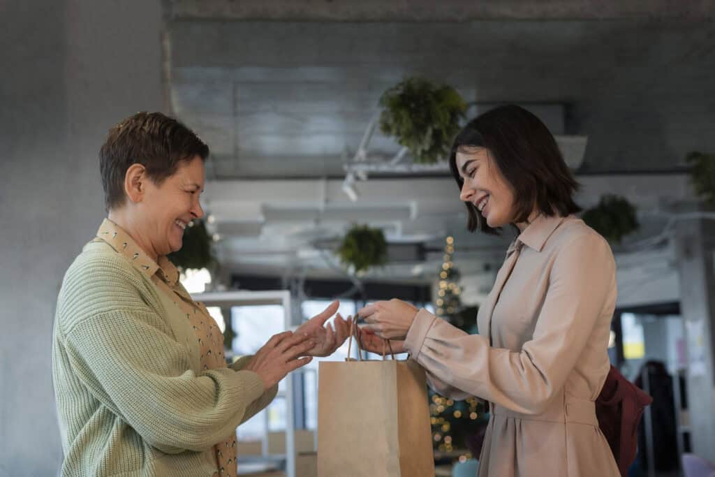 Two women smiling and exchanging a gift bag indoors, with festive decorations in the background. | Tsunami Reach