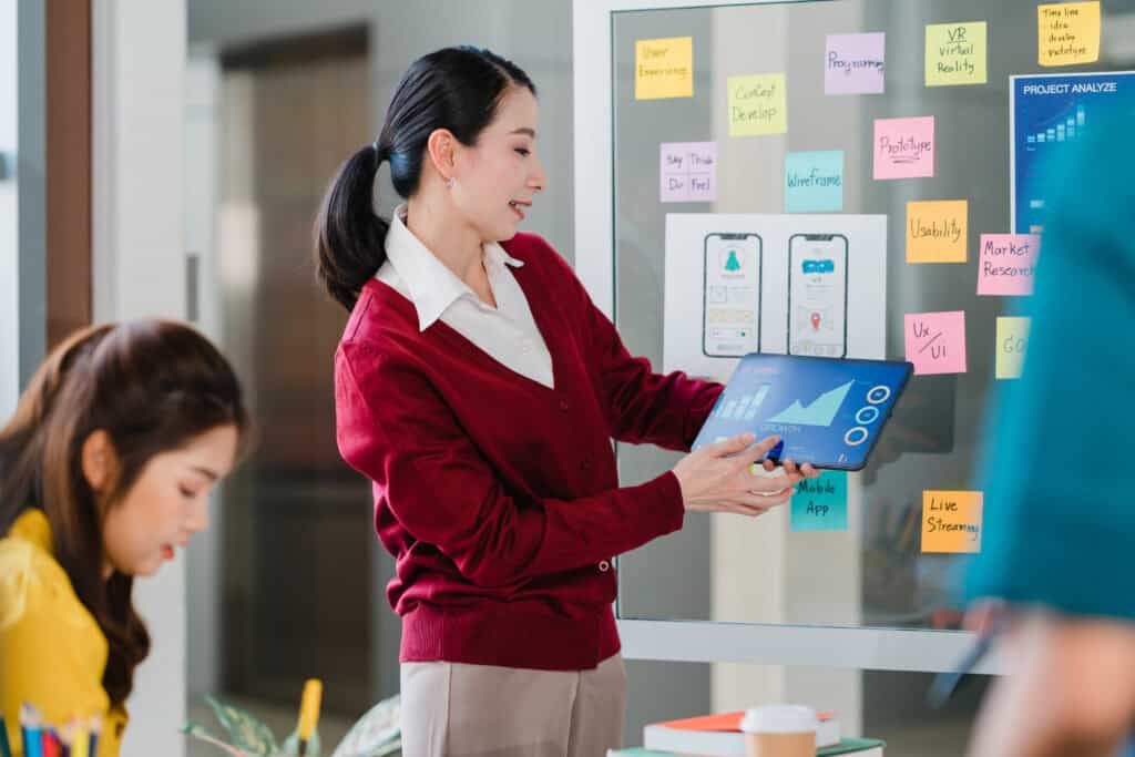 Woman in red cardigan presenting a tablet with a graph to colleagues near a glass wall covered in colorful sticky notes. | Tsunami Reach