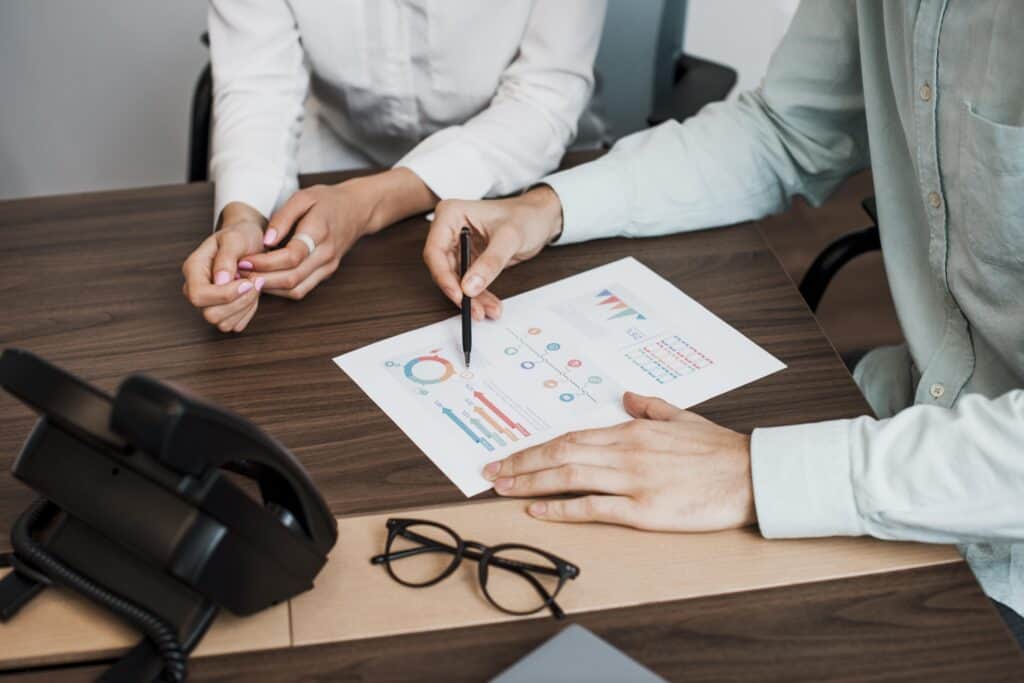 Two people discussing colorful charts and graphs on paper at a desk with a phone and glasses nearby. | Tsunami Reach