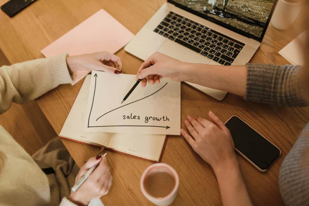 Two people discuss a sales growth chart at a table with a laptop, phone, notebook, and coffee mug. | Tsunami Reach