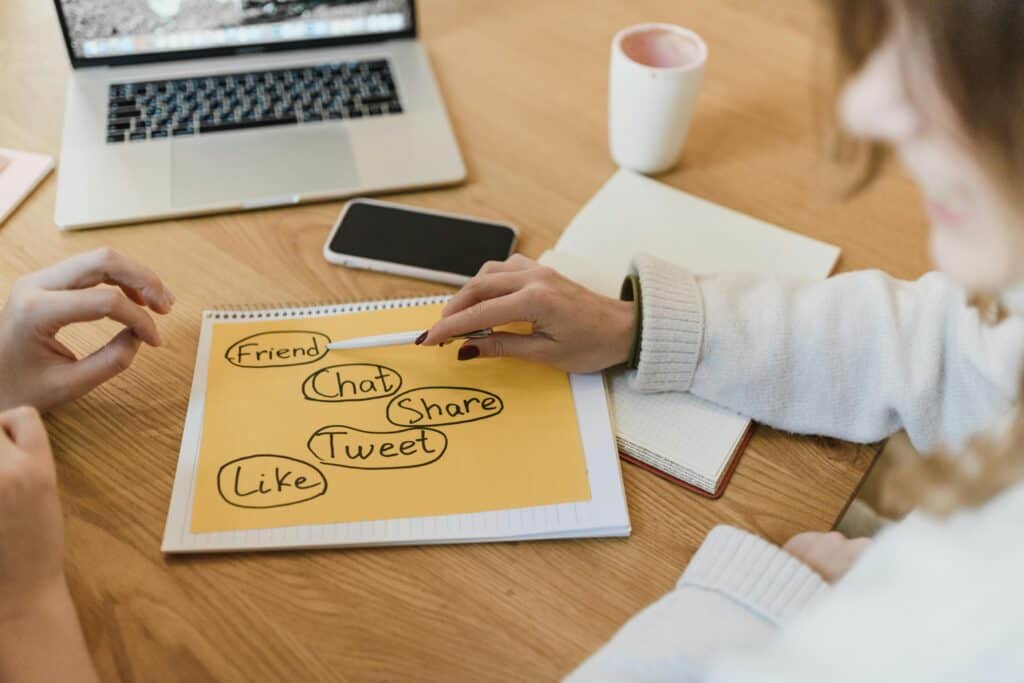 Two people brainstorming on social media terms written on paper near a laptop, phone, and coffee cup on a desk. | Tsunami Reach