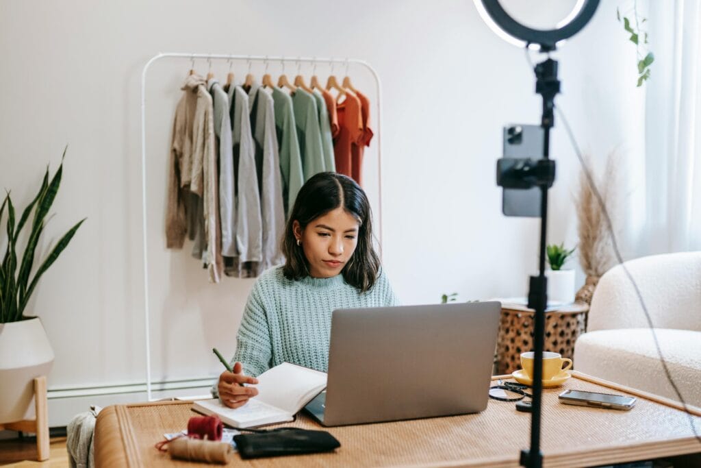 Woman working at a laptop with a notebook; clothes rack and camera setup in a bright, modern room. | Tsunami Reach