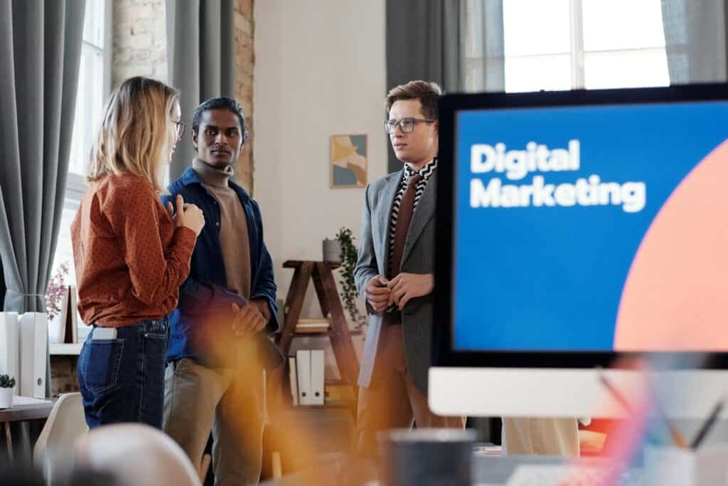 Three people have a discussion in an office, with a computer screen displaying "Digital Marketing" in the foreground. | Tsunami Reach