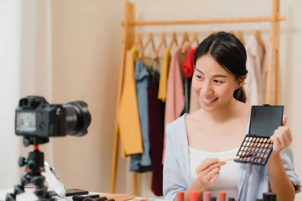 Smiling woman recording a makeup tutorial, holding an eyeshadow palette in front of a camera with clothes in the background. | Tsunami Reach
