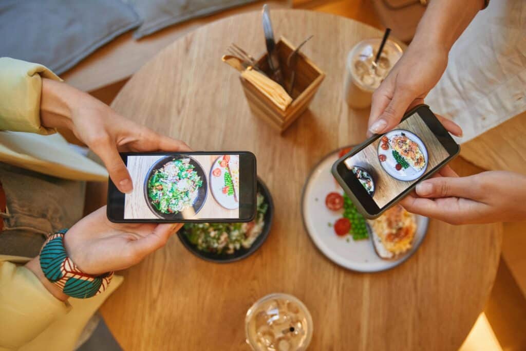 Two people take photos of their food with smartphones at a table set with salads, drinks, and cutlery. | Tsunami Reach